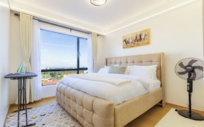 Ein Hotelzimmer mit einem großen Bett, einem Standventilator, einem Bügelbrett und Blick auf die Stadt.