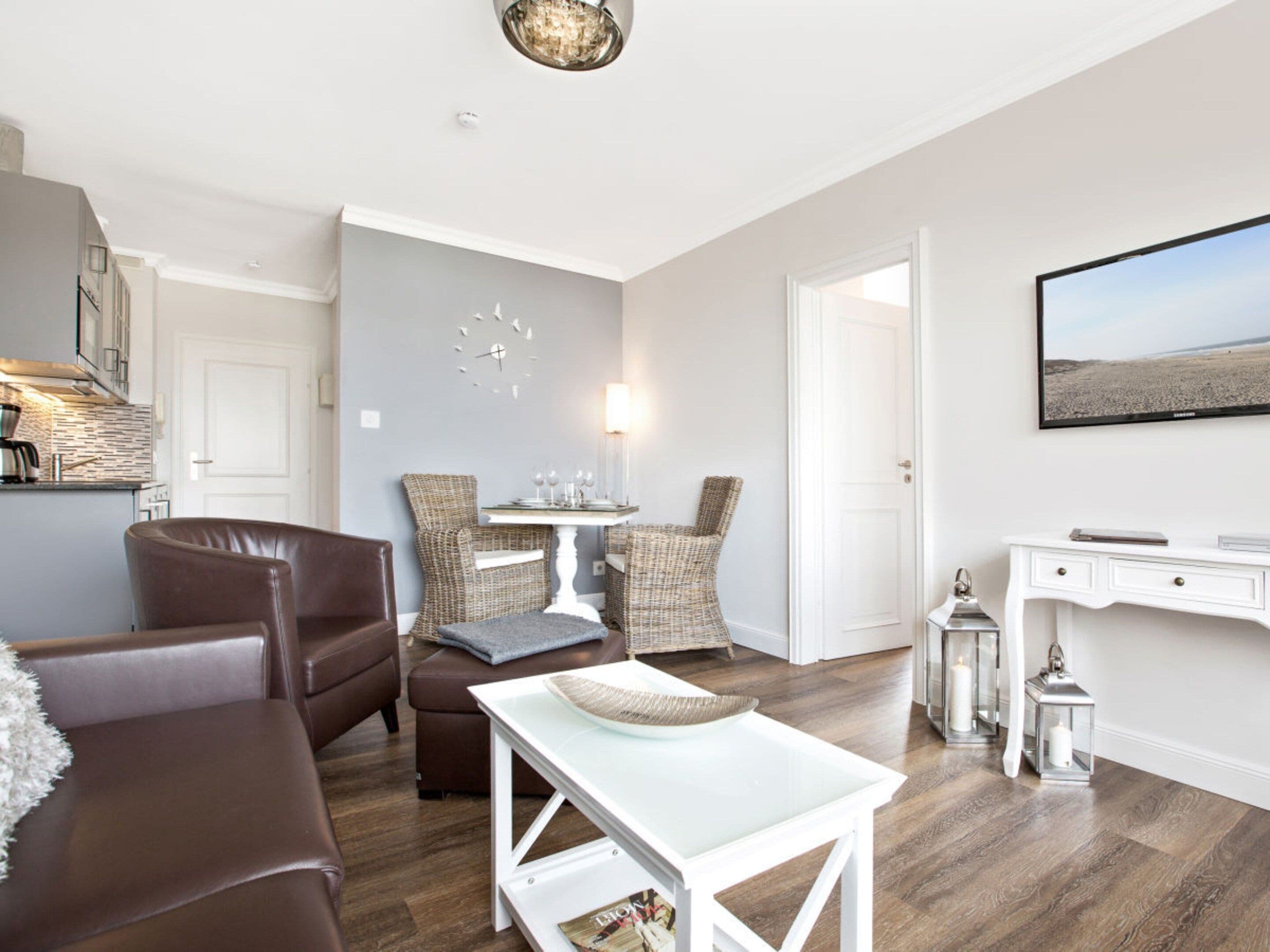 A modern living room with a brown leather sofa, a white coffee table, wicker chairs, a dining table, and a flat-screen TV mounted on the wall.