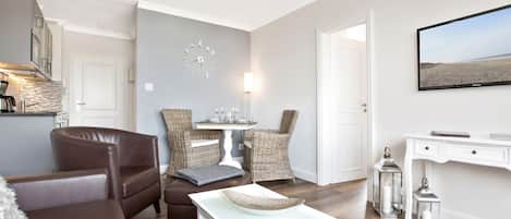 A modern living room with a brown leather sofa, a white coffee table, wicker chairs, a dining table, and a flat-screen TV mounted on the wall.