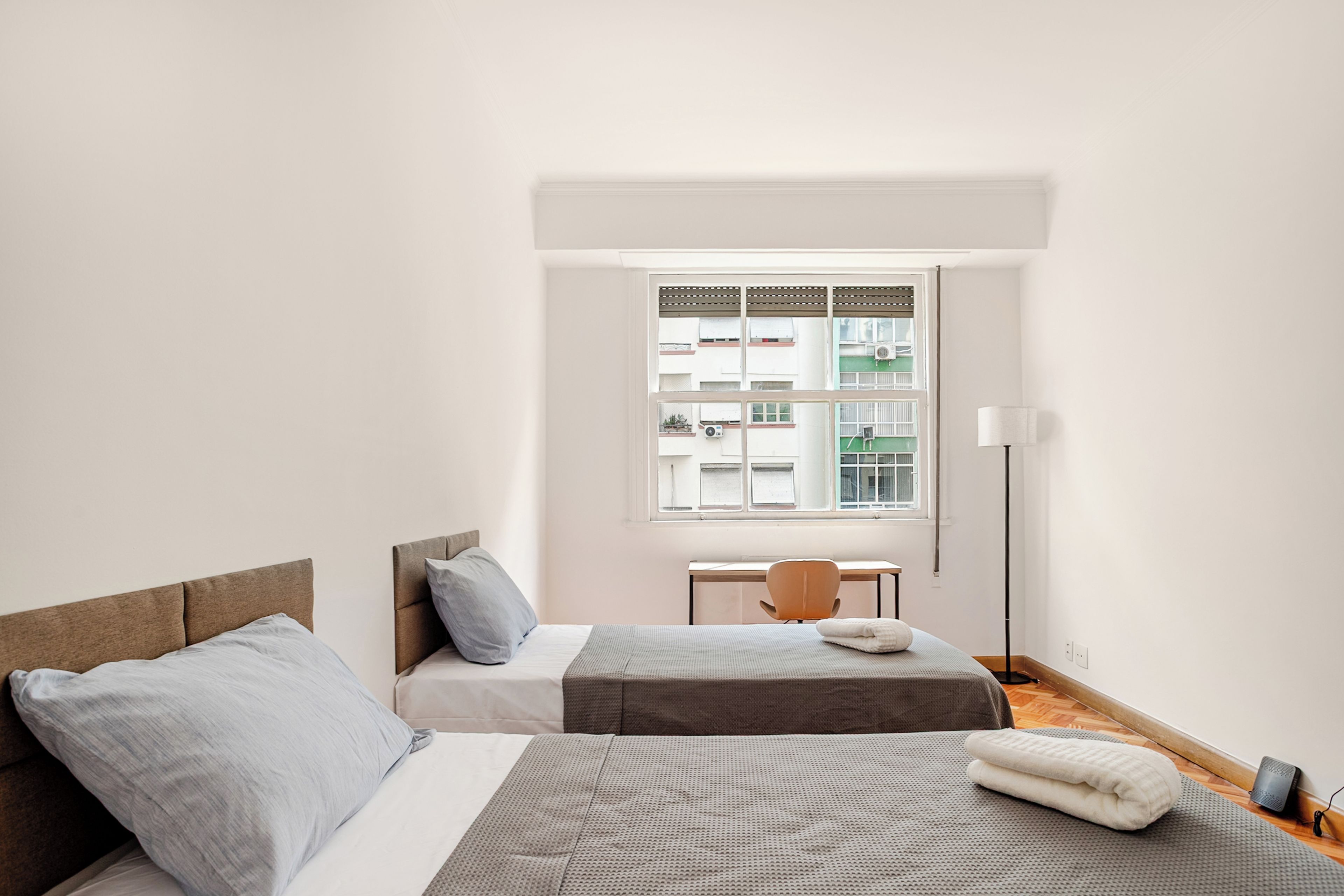 Une chambre avec deux lits, un bureau, une chaise, un lampadaire et une fenêtre donnant sur des immeubles.
