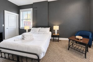 A hotel room with a bed, two bedside lamps, a blue chair, a small table, and a window with blinds.