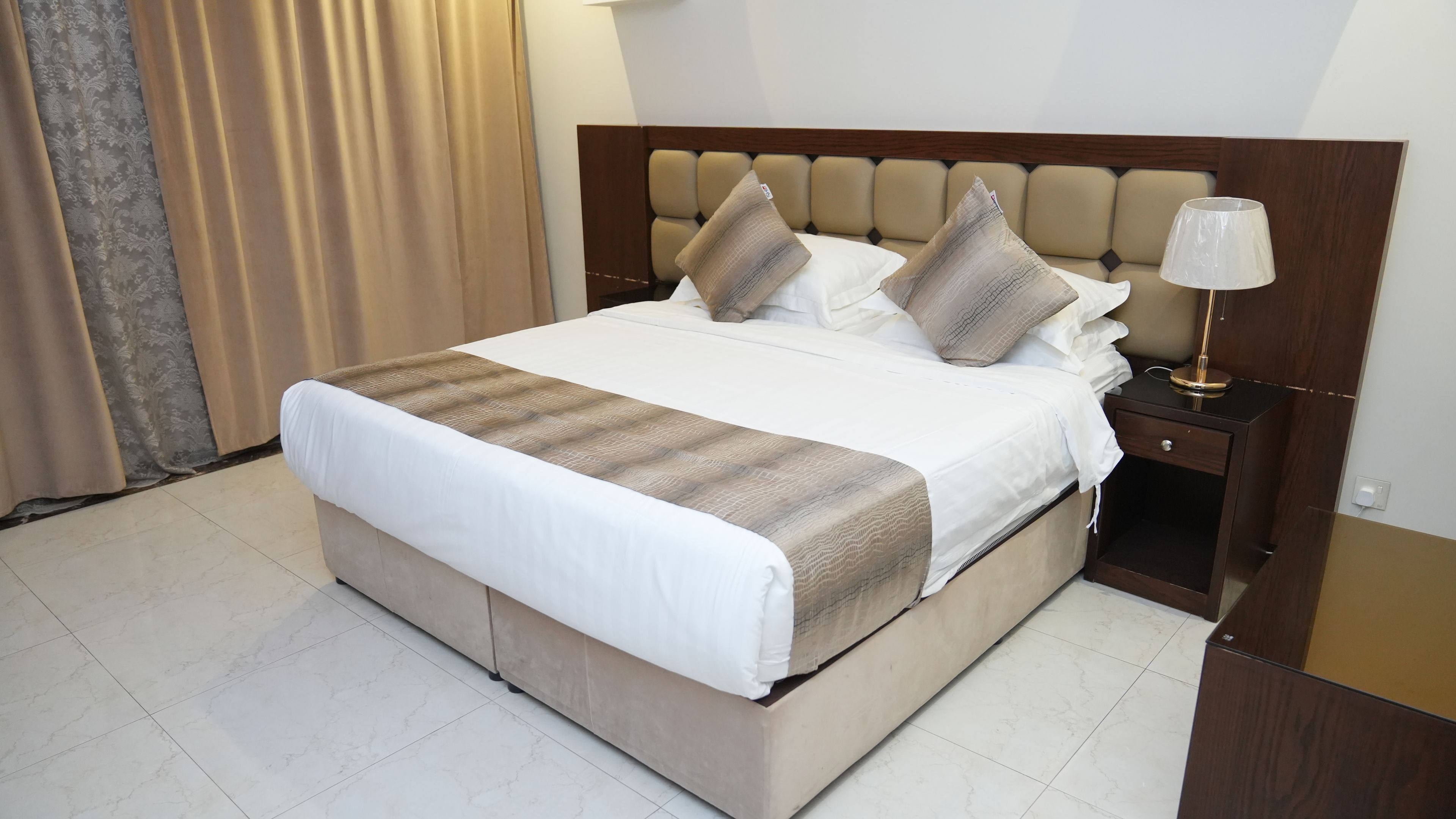 Ein Hotelzimmer mit Bett, Nachttisch, Lampe und Vorhang.