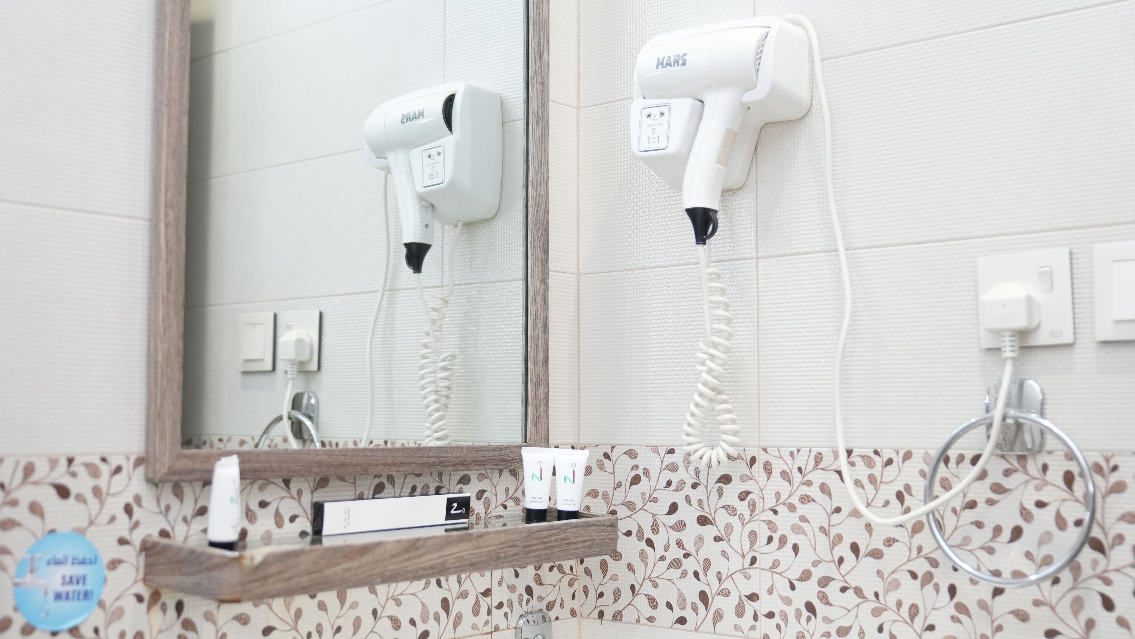 A bathroom with a mirror, two hair dryers mounted on the wall, a wooden shelf with toiletries, and a patterned tile backsplash.