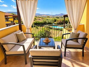 A balcony with a sofa and chairs, a table with a decorative item, and a view of a pool and landscape. - New Listing (Sardegna)