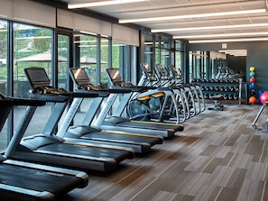 Ein Fitnessraum mit mehreren Laufbändern, die an einer Wand mit großen Fenstern aufgestellt sind, einem Hantelständer und Trainingsbällen.