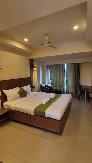 A hotel room with a large bed, a desk, a chair, a ceiling fan, and a window with curtains. - Tidemont by Velanora (Colva)