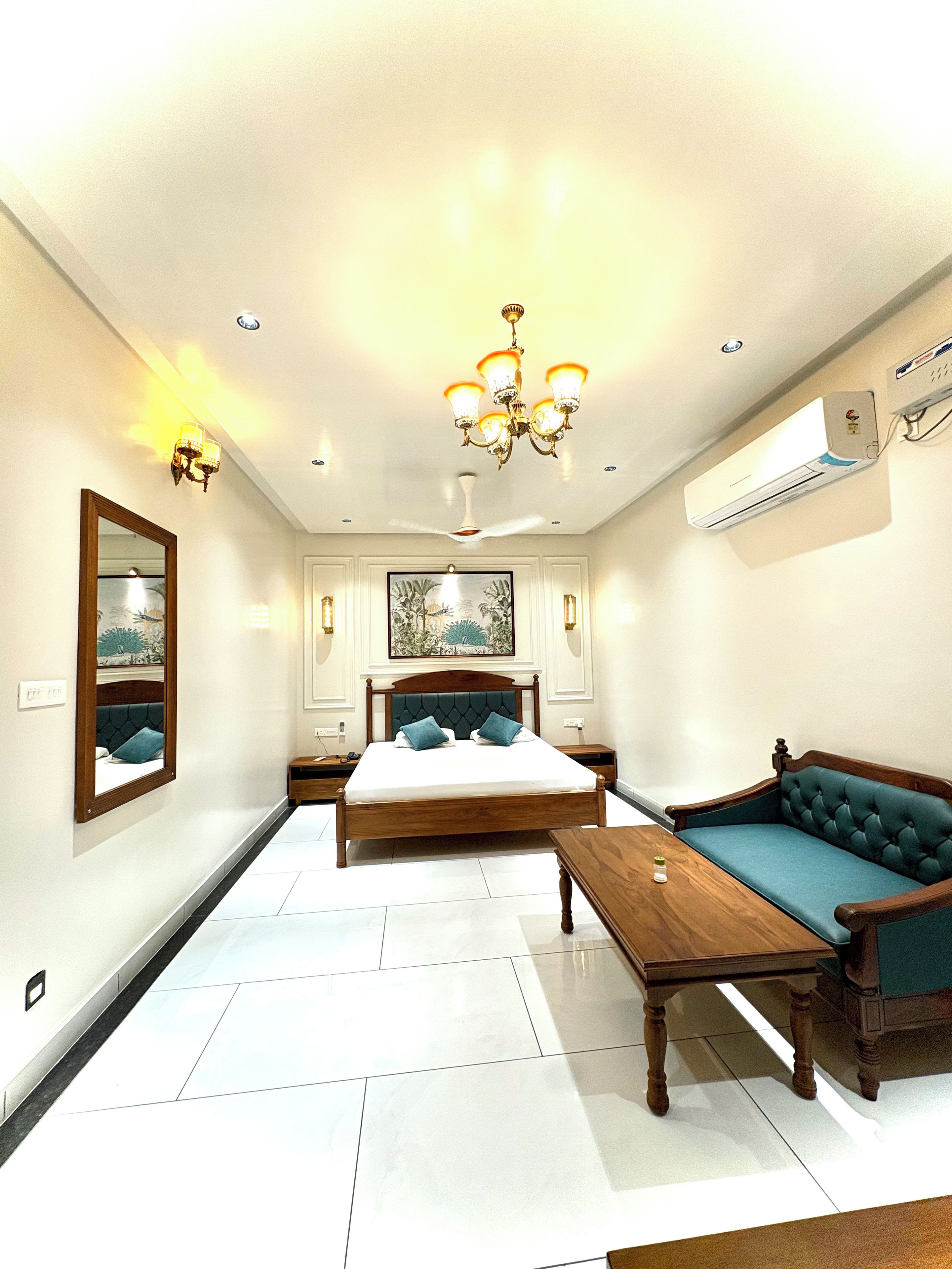 A bedroom with a bed, a sofa, a wooden table, a mirror, and a ceiling fan.