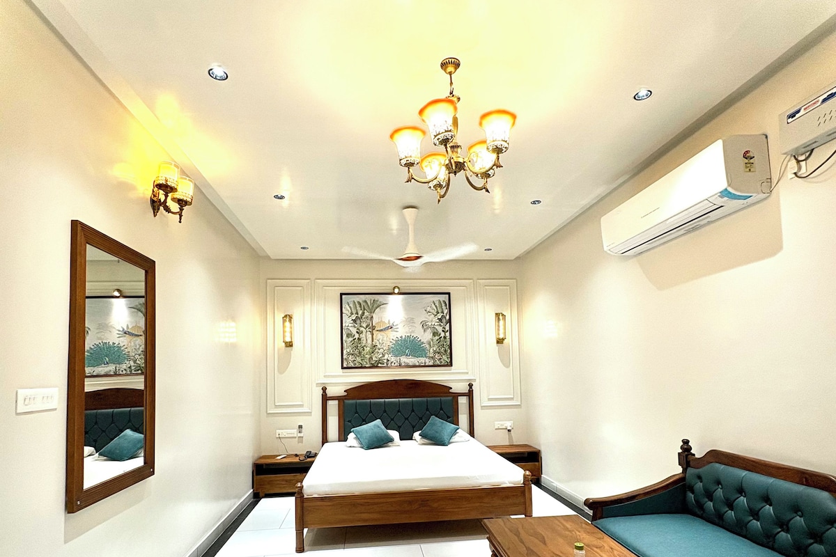 A bedroom with a bed, a sofa, a wooden table, a mirror, and a ceiling fan.