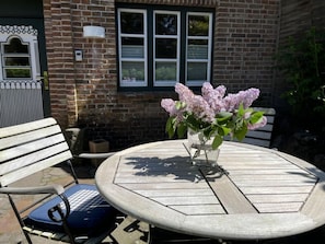 A round wooden table with a vase of flowers, a bench, and a brick building with windows in the background. - Holiday Apartment in Sylt With 1 Bedroom (Sylt)