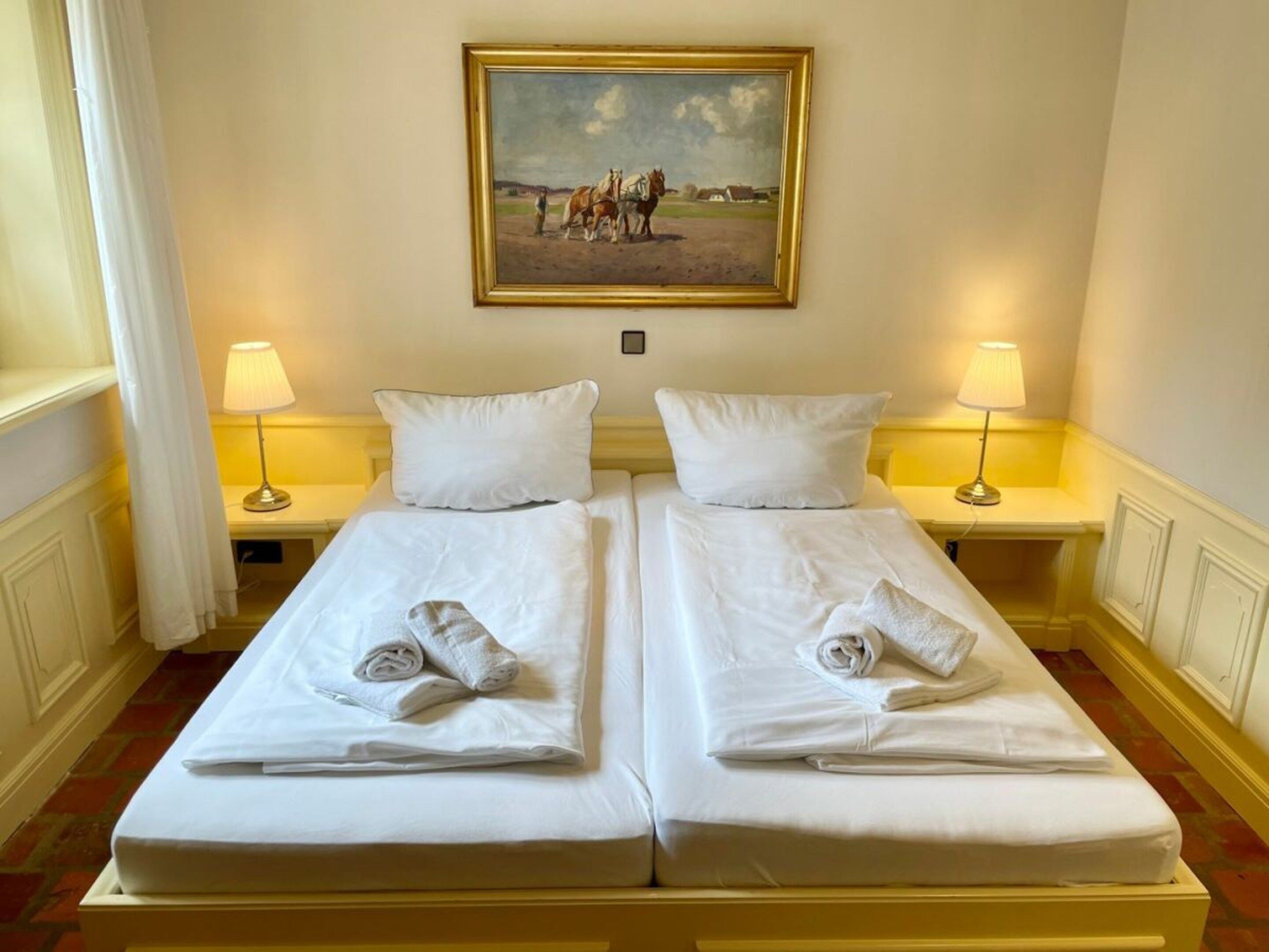 Una habitación de hotel con dos camas individuales, ropa de cama blanca, un cuadro de caballos en la pared y lámparas de noche.