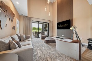 A modern living room with a large flat-screen TV, a sofa, a coffee table, and a view of the outdoors through a glass door. - Modern Oasis 4 BD/4.5 BA Magic Village by Pininfarina - 5501 TR (Kissimmee)