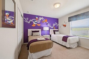 Quarto de hotel com duas camas, um mural na parede roxa, uma janela com persianas e uma luminária de teto.
