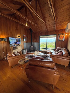 A cozy cabin interior with wooden walls, a fireplace, leather sofas, and a television. - Relax at our Beach Front Cosy 2 Bed Bach (Waitārere Beach)