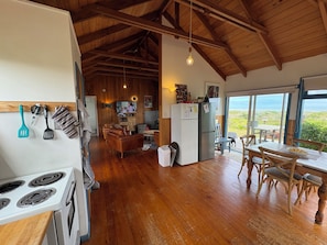 A spacious kitchen with wooden flooring, a dining area with wooden chairs, a refrigerator, and a television in the background. - Relax at our Beach Front Cosy 2 Bed Bach (Waitārere Beach)