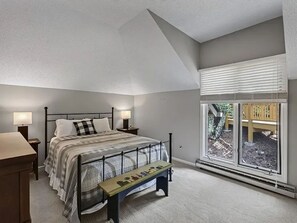 A bedroom with a sloped ceiling, a bed with a headboard, bedside lamps, a bench with a picture, and a large window with blinds. - Blue Ridge Mountain Retreat Overlooking the 18th Hole (Wintergreen Resort)