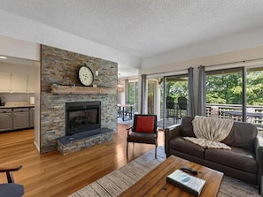 A modern living room with a stone fireplace, a leather sofa, and a dining area visible through large windows. - Blue Ridge Mountain Retreat Overlooking the 18th Hole (Wintergreen Resort)