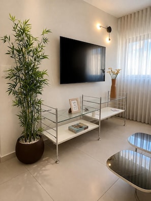 A modern living room with a large flat-screen TV mounted on the wall, a glass coffee table, a potted bamboo plant, and a minimalist shelving unit. - סוויטה מרווחת  וממוזגת מיקום מנצח 5דקות הליכה מהים במתחם בריכת שחייה 
sea said92 (Eilat)