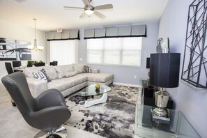 A modern living room with a sectional sofa, a glass coffee table, a black office chair, a ceiling fan, and a large window with blinds. - Cozy 4 BD/ 3.5 BA/ Compass Bay - 5149 AD (Kissimmee)