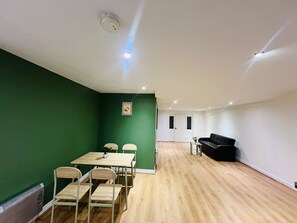 Ein Zimmer mit grüner Wand, Holzboden, einem Esstisch mit Stühlen, einer schwarzen Couch und einem kleinen Tisch mit einer Vase.