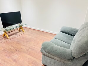 A living room with a flat-screen TV on a glass table with wooden legs, a grey sofa, and wooden flooring. - Extra-Large Manchester City Centre Flat | 2 Bedrooms, 2 Bathrooms & TV Room (Manchester)