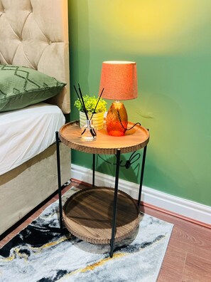 A round wooden side table with a lamp, a vase with plants, and a candle on a patterned rug in a room with a green wall and a tufted headboard. - Extra-Large Manchester City Centre Flat | 2 Bedrooms, 2 Bathrooms & TV Room (Manchester)