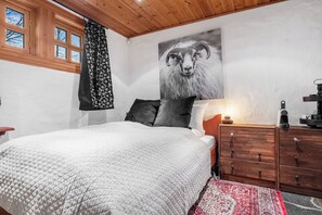 Un dormitorio con cama, cómoda de madera, mesita de noche, lámpara de pared y un cuadro grande con la imagen de un carnero.