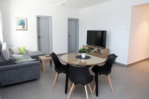 A modern living room with a sofa, a glass-top dining table with chairs, a flat-screen TV, and a small potted plant. - Lity Apartment Green (Podglavica)