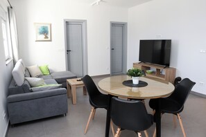A modern living room with a grey sofa, a round glass table, black chairs, a flat-screen TV, and a small potted plant. - Lity Apartment Green (Podglavica)