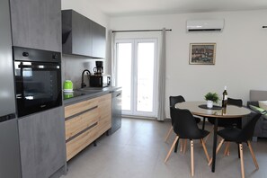 A modern kitchen with a built-in oven, a coffee maker, and a dining area with a round table and chairs. - Lity Apartment Green (Podglavica)