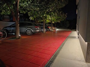 A nighttime parking area with red-tiled flooring, parked cars, and trees providing shade. - Lity Apartment Green (Podglavica)