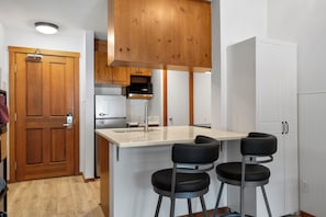 A compact kitchen with a wooden door, a built-in microwave, a refrigerator, and a bar area with stools. - Ski-In/Out Luxury Village Studio! (Sun Peaks)