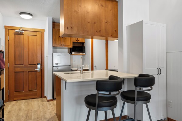 A compact kitchen with a wooden door, a built-in microwave, a refrigerator, and a bar area with stools. - Ski-In/Out Luxury Village Studio! (Sun Peaks)