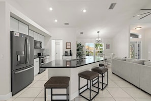 A modern kitchen with a central island, bar stools, and a grey sofa in the adjacent living area. - The Perfect 3BR Villa | Beautiful Lanai and Luxury (Cape Coral)