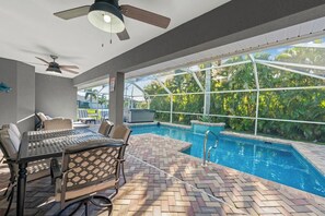 An indoor swimming pool area with a table and chairs, ceiling fans, and a view of lush greenery outside. - The Perfect 3BR Villa | Beautiful Lanai and Luxury (Cape Coral)