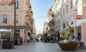 A pedestrian street lined with cafes and shops, with outdoor seating and a large planter with shrubs. - New Listing (Saint-Raphaël)