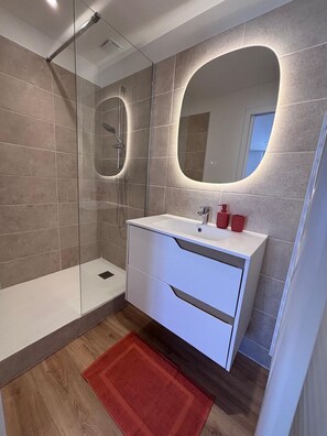 A modern bathroom with a glass-enclosed shower, a white vanity with a mirror, and a red rug on the floor. - New Listing (Saint-Raphaël)