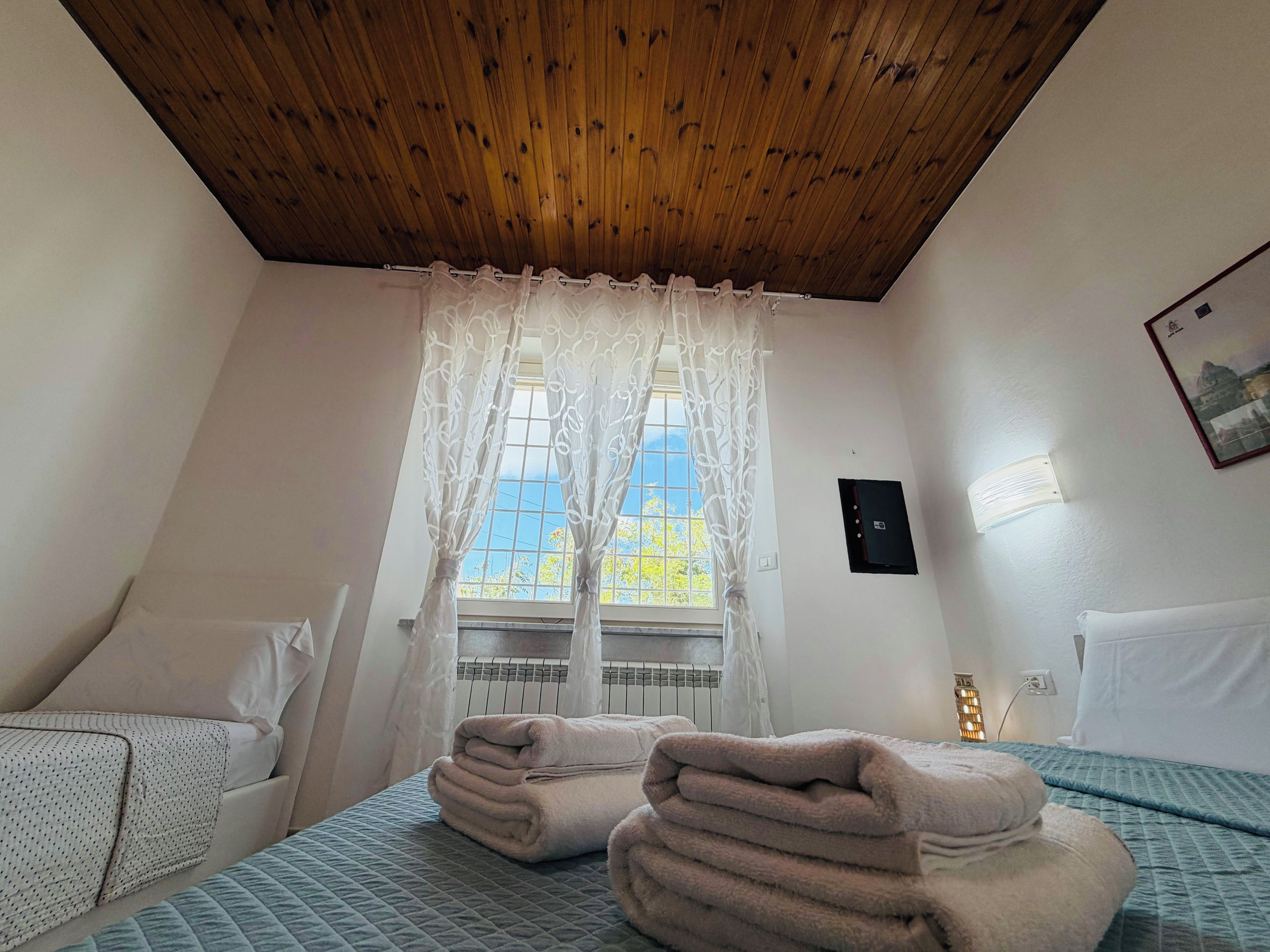 A hotel room with a wooden ceiling, a window with sheer curtains, a bed with a quilted bedspread, and a bedside table with a pillow.