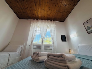 Une chambre d’hôtel avec un plafond en bois, une fenêtre ornée de rideaux légers, un lit recouvert d’une courtepointe matelassée et une table de chevet avec un oreiller.