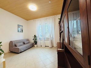 Una habitación con techo de madera, un sofá gris, una planta en maceta, una ventana con cortinas y un armario.