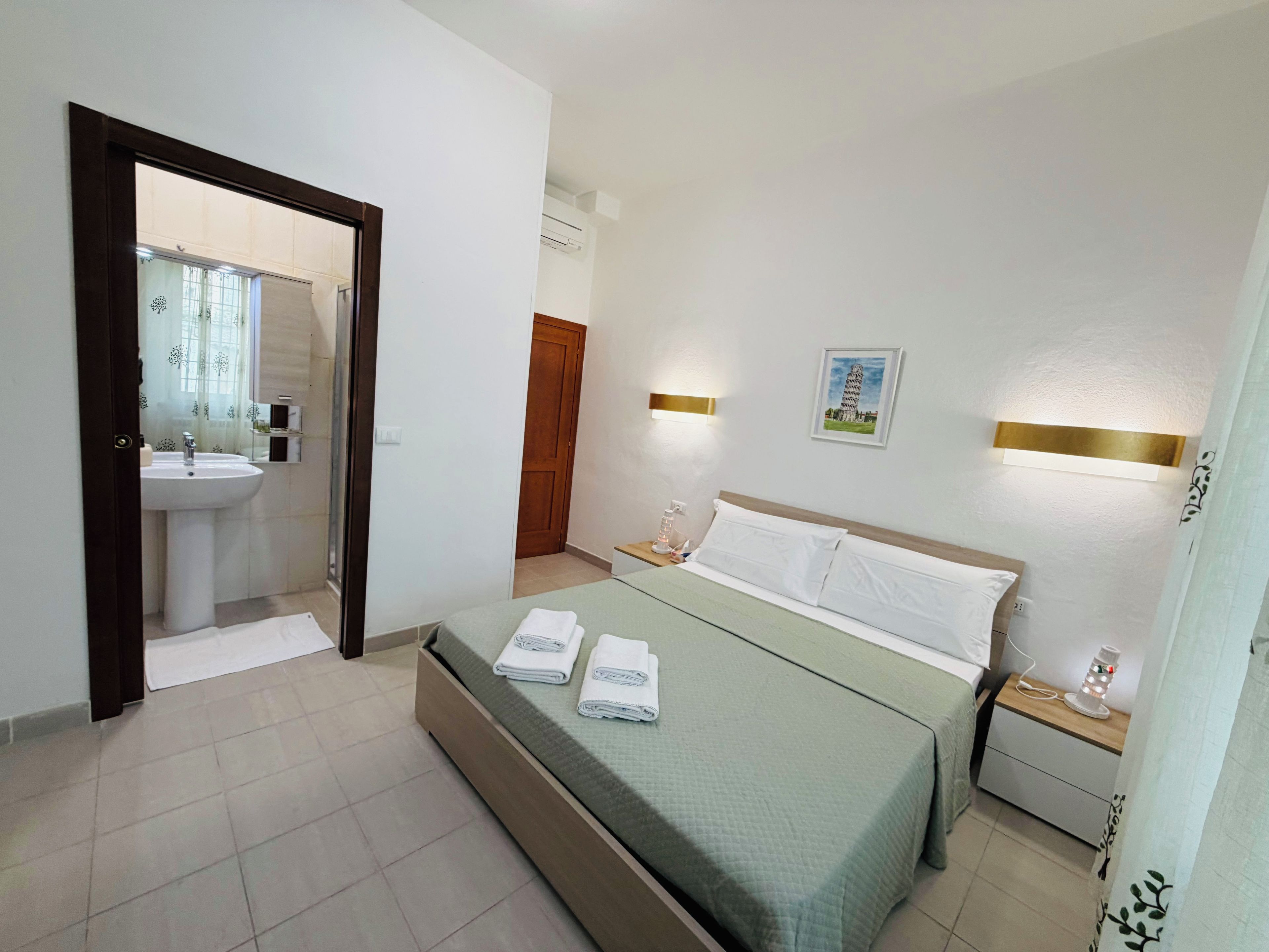 Habitación de hotel con cama, mesitas de noche, lámpara de pared, un cuadro de la Torre inclinada de Pisa y un baño visible a través de una puerta abierta.