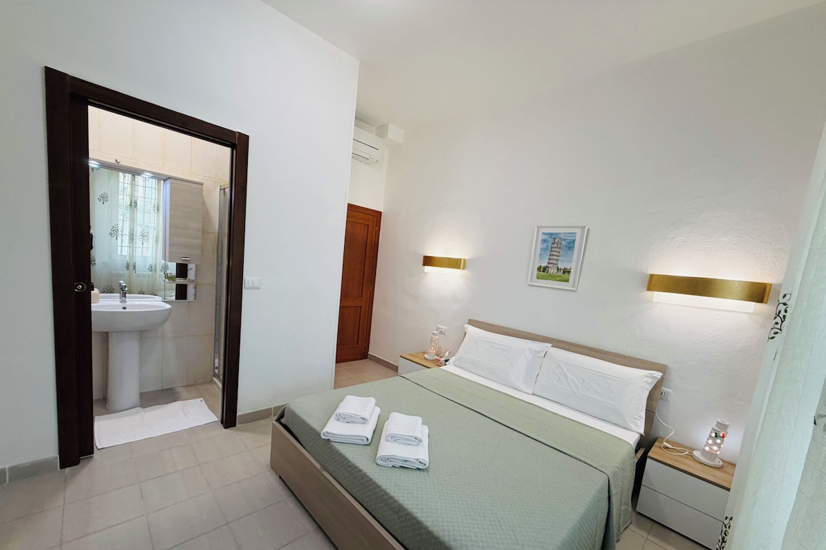 Habitación de hotel con cama, mesitas de noche, lámpara de pared, un cuadro de la Torre inclinada de Pisa y un baño visible a través de una puerta abierta.