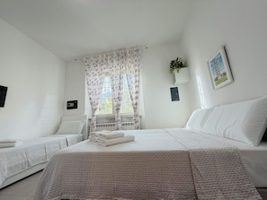 Habitación de hotel con dos camas, una ventana con cortinas florales, una planta en la pared y un cuadro enmarcado.