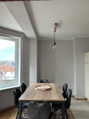 A dining area with a wooden table, black chairs, and a hanging light fixture. - New Listing (De Panne)