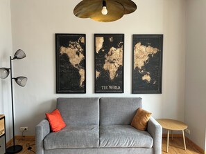 A grey sofa with two cushions, a floor lamp, and three world map posters on the wall. - New Listing (De Panne)