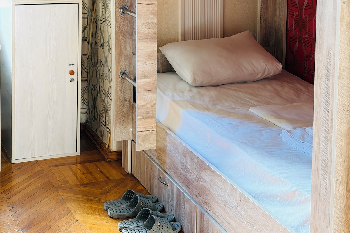 A bunk bed with a wooden headboard, a single bed with a pillow, and a pair of slippers on the floor.