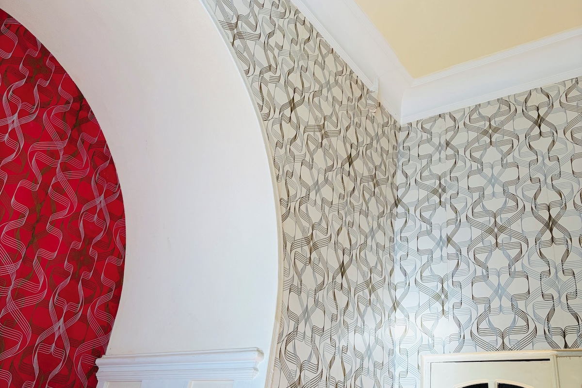 A room with a curved wall, a bed with white bedding, a patterned wallpaper, and a window with frosted glass.