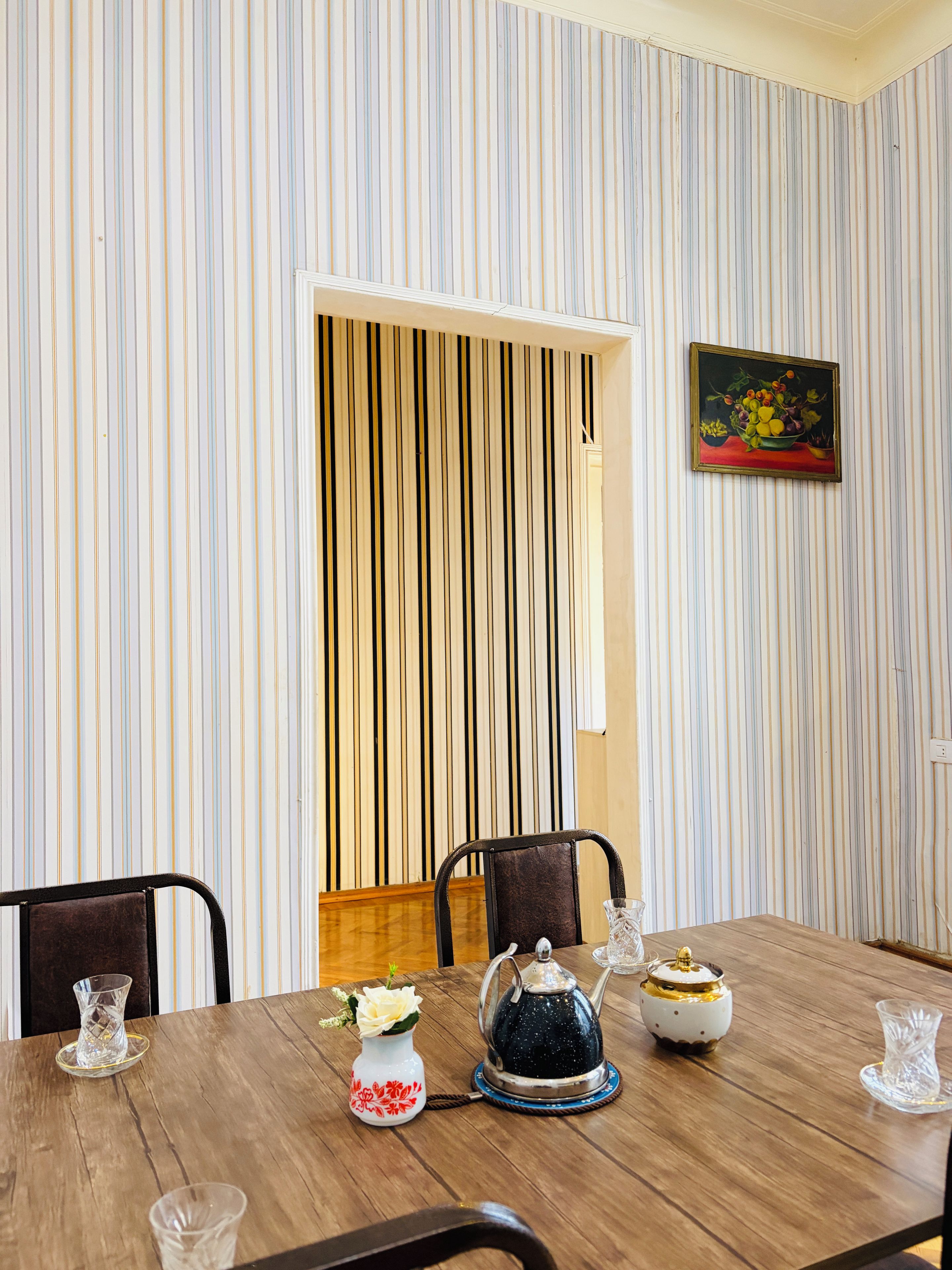 Een eethoek met gestreept behang, een lijstwerk schilderij, en een houten tafel gedekt met een theepot, een vaas met bloemen, en een glas.