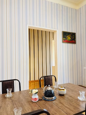 A dining area with a striped wallpaper, a framed painting, and a wooden table set with a teapot, a vase with flowers, and a glass. - Alina Central Hostel (Baku)