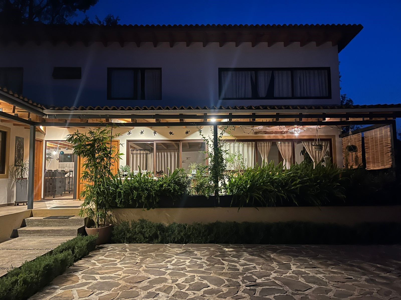 A two-story house with a stone patio, a covered outdoor seating area, and a garden with potted plants.