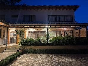 A two-story house with a stone patio, a covered outdoor seating area, and a garden with potted plants.